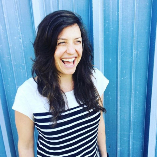 Black-haired light-skinned woman laughing against a bright blue background.